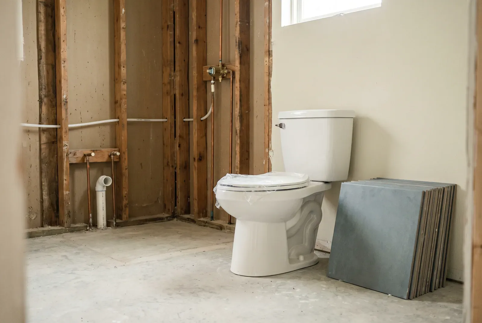 A bathroom partway through a remodel, with open stud walls, a roughed-in shower valve and supply lines, a new toilet and a stack of floor tile.